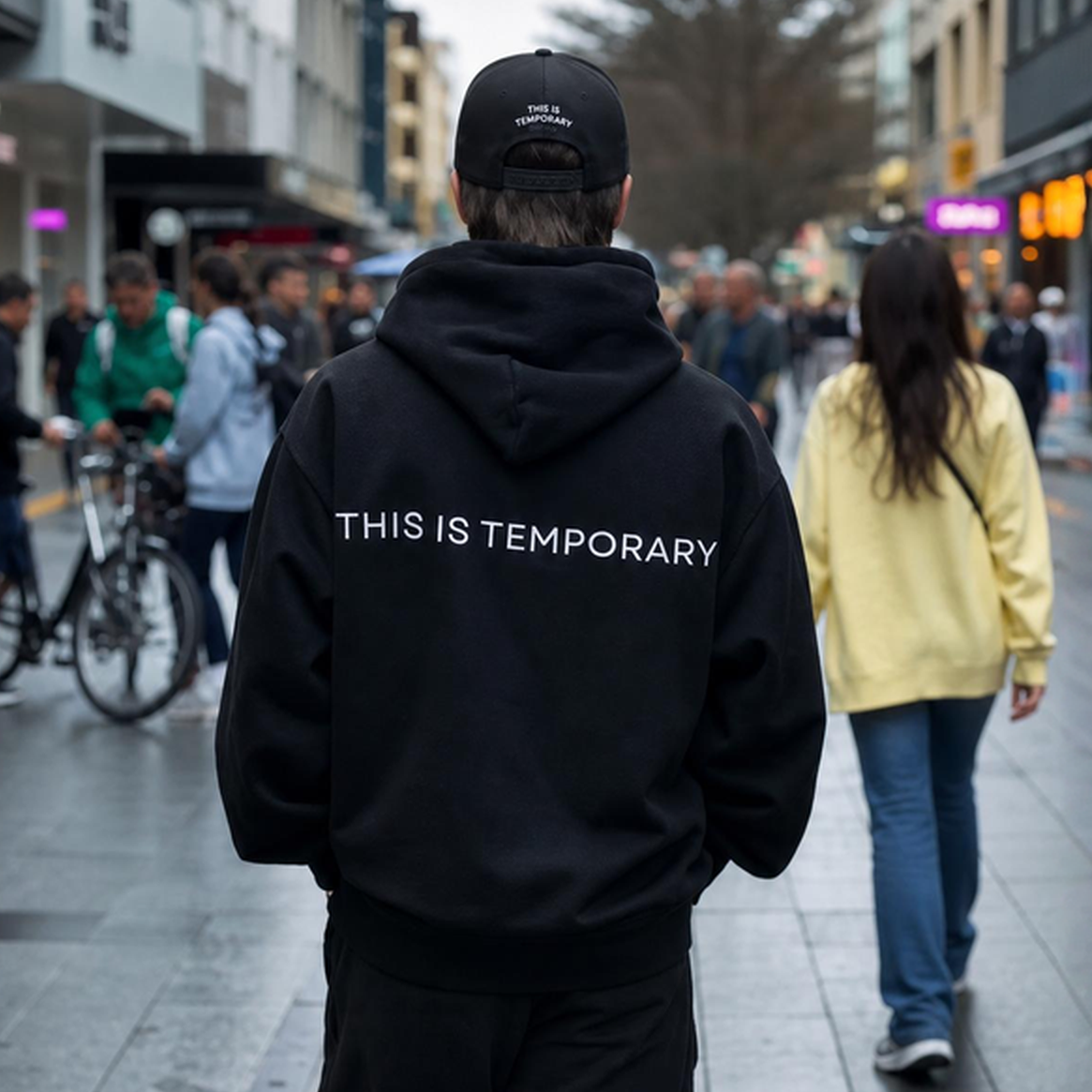 Rear view of a model wearing the TMPRY Initiation Hood in 400GSM raw black fleece, featuring the rear "THIS IS TEMPORARY" lettering and architectural drape.