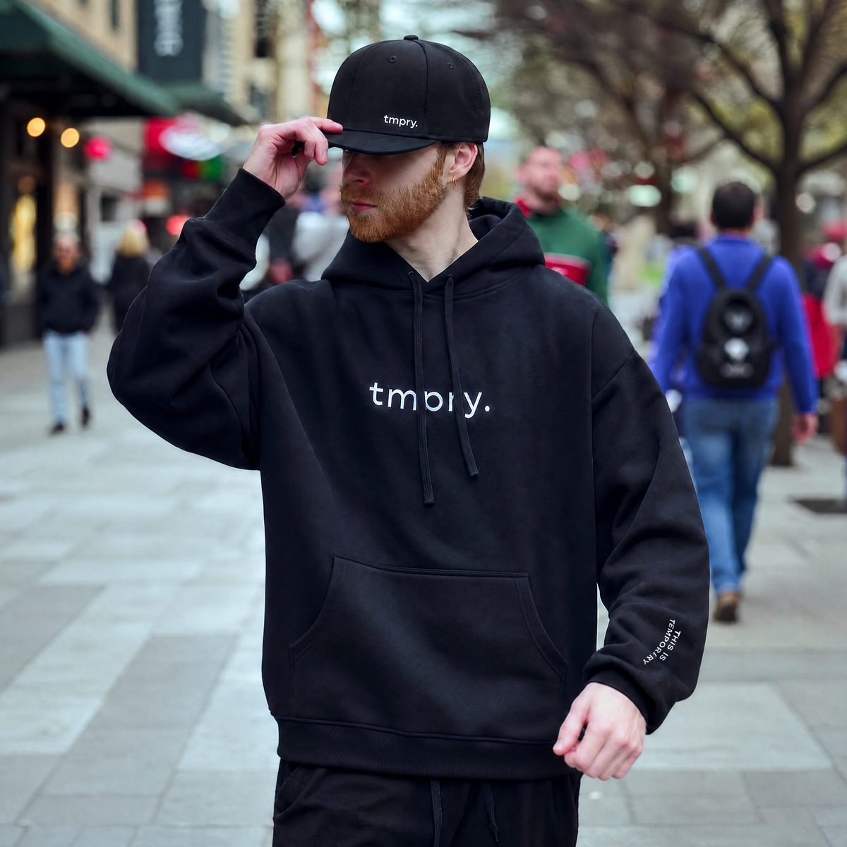 Front view of a model wearing the TMPRY Initiation Hood in 400GSM raw black fleece, featuring the tmpry. logo and "THIS IS TEMPORARY" sleeve branding.